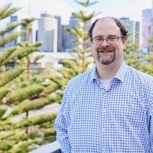 An image of Steven, a man with glasses and a blue shirt. he is smiling. there is a norfolk pine tree and a cityscape in the background.